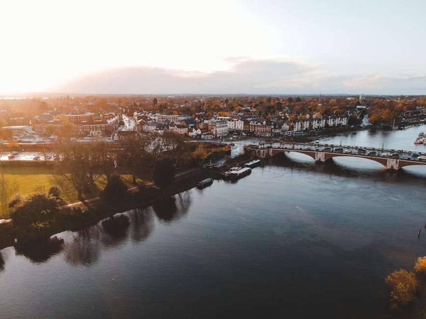 An aerial view of Kingston's riverside, taken during the golden hour, showcasing a gently flowing river bordered by lush trees and well-maintained garden beds. In the background, a variety of resident