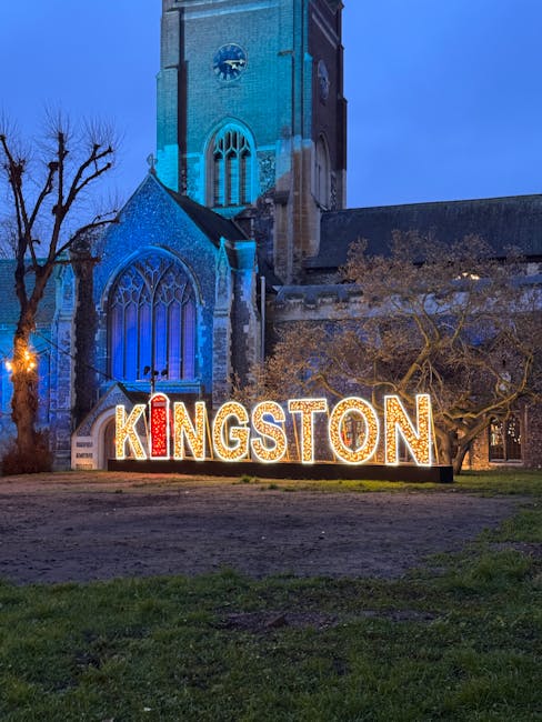 A nighttime view of a historic church with a tall, stone clock tower and large arched stained-glass windows illuminated by blue lighting. In the foreground, there is a landscaped garden area with neat